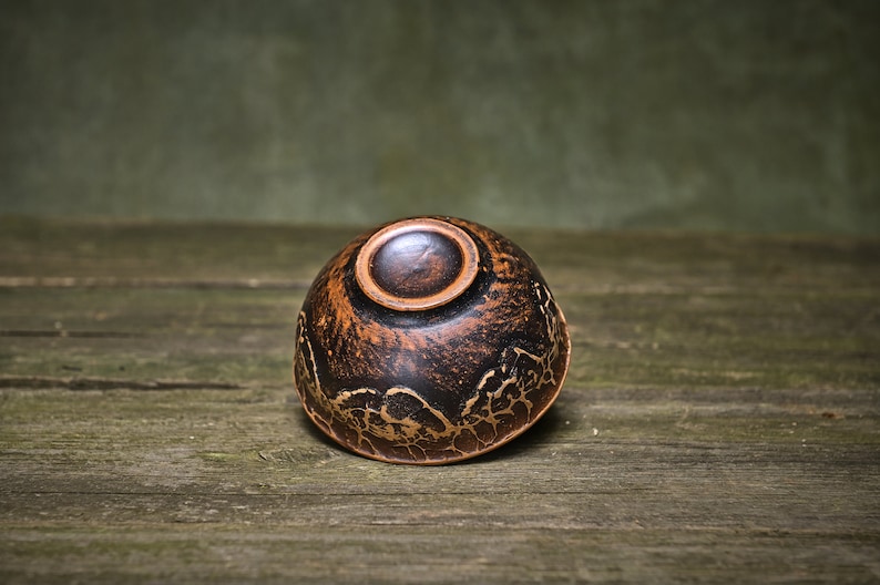 May include: A small, dark brown ceramic bowl with a gold-colored design. The bowl is sitting on a wooden surface.