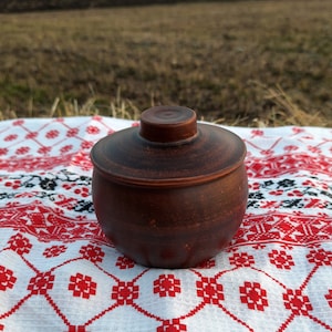 Könnte beinhalten: Ein kleiner, runder, brauner Keramiktopf mit Deckel. Der Topf steht auf einem weißen Tuch mit einem roten und schwarzen Blumenmuster. Der Hintergrund ist ein Feld unter einem bewölkten Himmel.