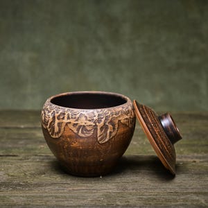 Peut inclure: Un bol en céramique marron fait à la main avec un couvercle. Le bol présente un motif texturé et sculpté sur le bord supérieur. Le couvercle est légèrement ouvert, révélant l'intérieur sombre. Le bol repose sur une surface en bois vieilli.
