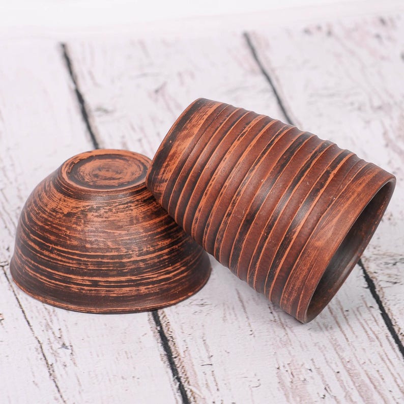 May include: A brown clay bowl and a matching cylindrical cup, both featuring a textured surface with horizontal ridges and a dark, mottled finish. The bowl is positioned on its side, while the cup stands upright next to it.