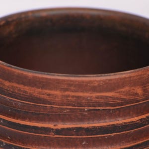 May include: A cylindrical clay pot with a dark reddish-brown finish. The exterior surface features horizontal ridges and visible brush-like texture marks throughout the body. The rim is thick and rounded, leading into a deep, dark interior cavity.