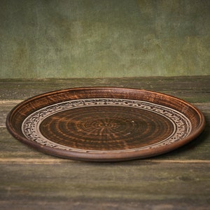 May include: A brown ceramic plate with a decorative design around the rim. The plate is sitting on a wooden surface.