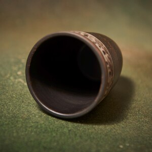 May include: A dark brown ceramic cup with a decorative band near the top. The cup is cylindrical with a wide opening and sits on a green surface. The interior of the cup is black.