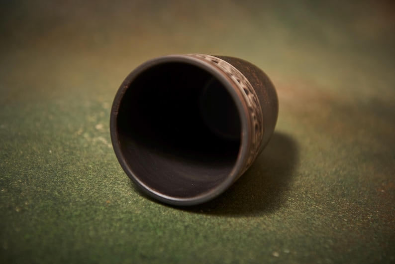 May include: A dark brown ceramic cup with a decorative band near the top. The cup is cylindrical with a wide opening and sits on a green surface. The interior of the cup is black.
