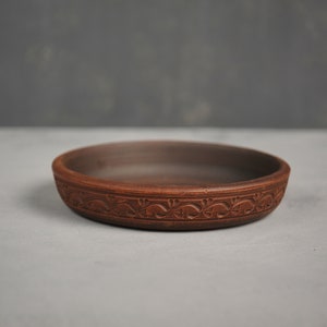 Könnte beinhalten: Eine braune Keramikschüssel mit einem dekorativen Muster am Rand. Die Schüssel ist leer und steht auf einer grauen Oberfläche.