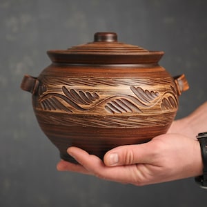 May include: A brown ceramic pot with a lid and handles. The pot has a carved design around the middle. The pot is held in a person's hands.