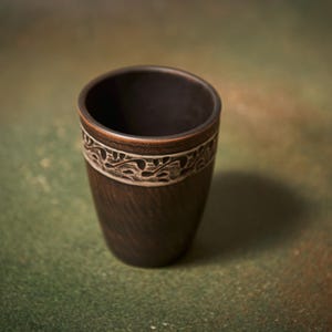 May include: A dark brown ceramic cup with a decorative band. The band features a white, carved floral design. The cup has a smooth, tapered shape and sits on a green and brown surface. The cup is approximately 4 inches tall.