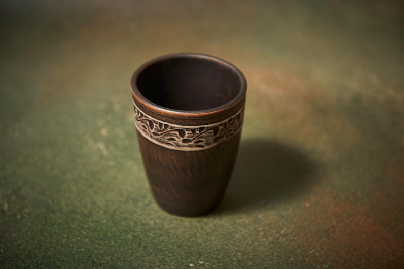 May include: A dark brown ceramic cup with a decorative band. The band features a white, carved floral design. The cup has a smooth, tapered shape and sits on a green and brown surface. The cup is approximately 4 inches tall.