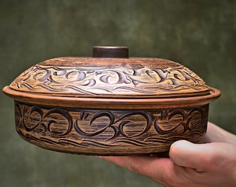 May include: A round brown clay casserole dish with a matching lid. The exterior of the pot and the lid feature dark brown carved swirling patterns and decorative motifs. The vessel has a smooth rim and a central knob on the lid.