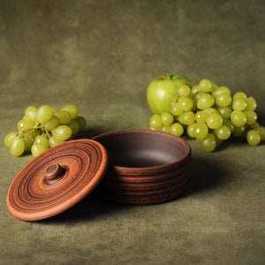 May include: A handmade, open, brown ceramic bowl with a matching lid, next to green grapes and a green apple. The bowl has a rustic, earthy appearance, suggesting it's suitable for serving or storage. The background is a muted green.
