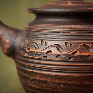May include: Close-up of a brown ceramic teapot with a lid. The teapot has a textured surface with carved floral patterns and horizontal lines. The spout and handle are visible, and the overall design is rustic.