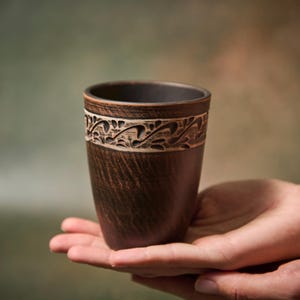 Peut inclure: Une tasse en céramique marron faite à la main avec une bande décorative de motifs floraux sculptés. La tasse est tenue dans des mains ouvertes, mettant en valeur sa texture et son design rustiques. La tasse mesure environ 10 cm de haut.