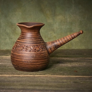 May include: A brown clay coffee pot with a wide, rounded body, a flared rim, and a long, tapered handle. The exterior features horizontal ridges and a decorative band of carved, repeating leaf-like patterns encircling the midsection of the vessel.