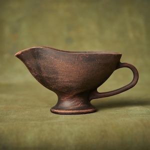May include: A brown ceramic gravy boat with a handle and a pedestal base. The boat is on a green background.