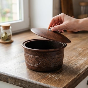 May include: A brown ceramic casserole dish with a lid being lifted. The dish has a decorative embossed pattern around the sides. The lid has a small knob. The dish is on a wooden surface.