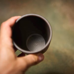 May include: A dark gray ceramic cup is held in a hand. The cup has a simple, rounded design and a smooth, matte finish. The interior of the cup is also dark gray.