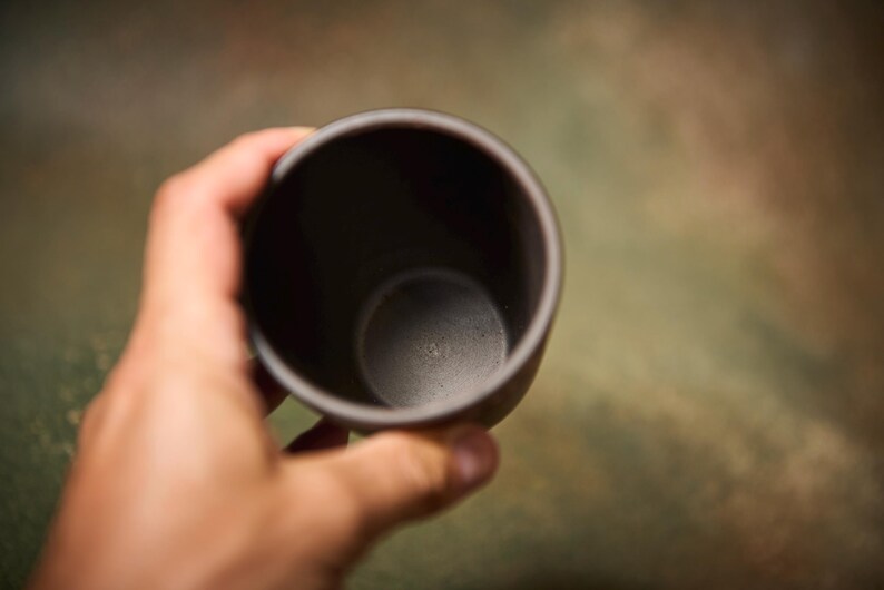 May include: A dark gray ceramic cup is held in a hand. The cup has a simple, rounded design and a smooth, matte finish. The interior of the cup is also dark gray.