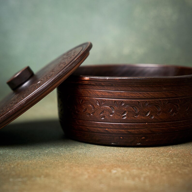 May include: A dark brown circular clay container with a matching lid. The exterior of the container and the top of the lid feature a carved, repeating vine-like pattern. The lid has a small, centered knob for lifting.