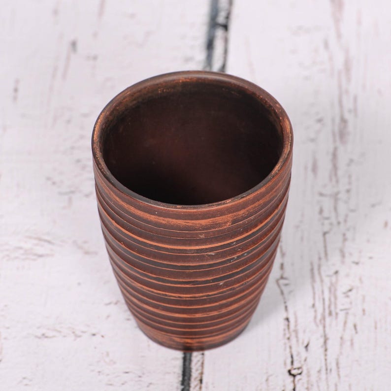 May include: A cylindrical brown clay cup featuring a series of horizontal, incised grooves circling the exterior. The vessel has a smooth, open top rim and a tapered base, showcasing the natural, earthy texture and deep reddish-brown color of the material.
