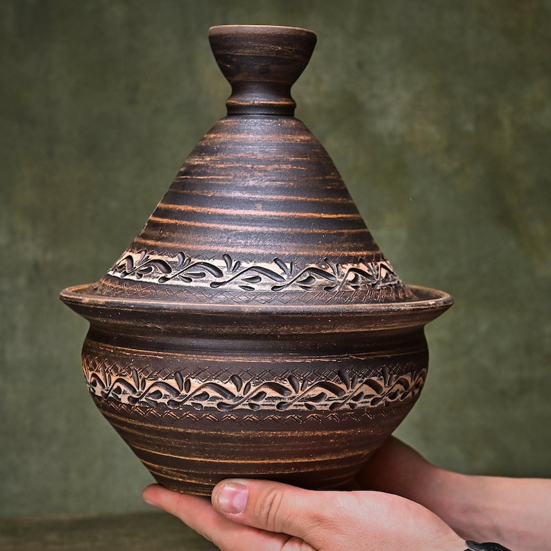 May include: A dark brown clay tagine with a conical lid and a wide base. The exterior features horizontal bands of carved leaf and vine patterns circling both the lid and the bowl. The surface has a natural, earthy texture with visible striations.
