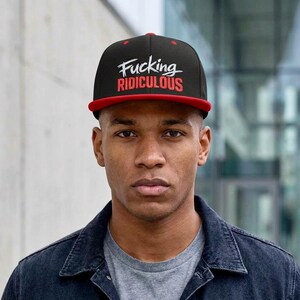 May include: Black and red baseball cap with the text "Fucking Ridiculous" in white and red. The cap has a flat brim and is worn by a person wearing a denim jacket and a gray t-shirt.