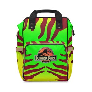 May include: A backpack featuring a vibrant neon green and yellow gradient background with dark red tiger-like stripes. The front pocket displays the circular Jurassic Park logo with a black dinosaur silhouette and the text Jurassic Park in yellow.
