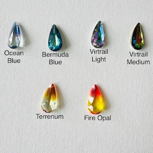 Op de afbeelding: Zes druppelvormige kristallen in verschillende kleuren worden getoond tegen een witte achtergrond. De kristallen zijn gelabeld: Ocean Blue, Bermuda Blue, Virtrail Light, Virtrail Medium, Terrenum en Fire Opal. Elk kristal heeft een facetontwerp.