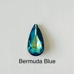 Op de afbeelding: Een druppelvormig Bermuda Blue kristal met een puntige top. Het kristal heeft een facetgeslepen oppervlak dat licht reflecteert, waardoor een glinsterend effect ontstaat. De tekst "Bermuda Blue" staat eronder.