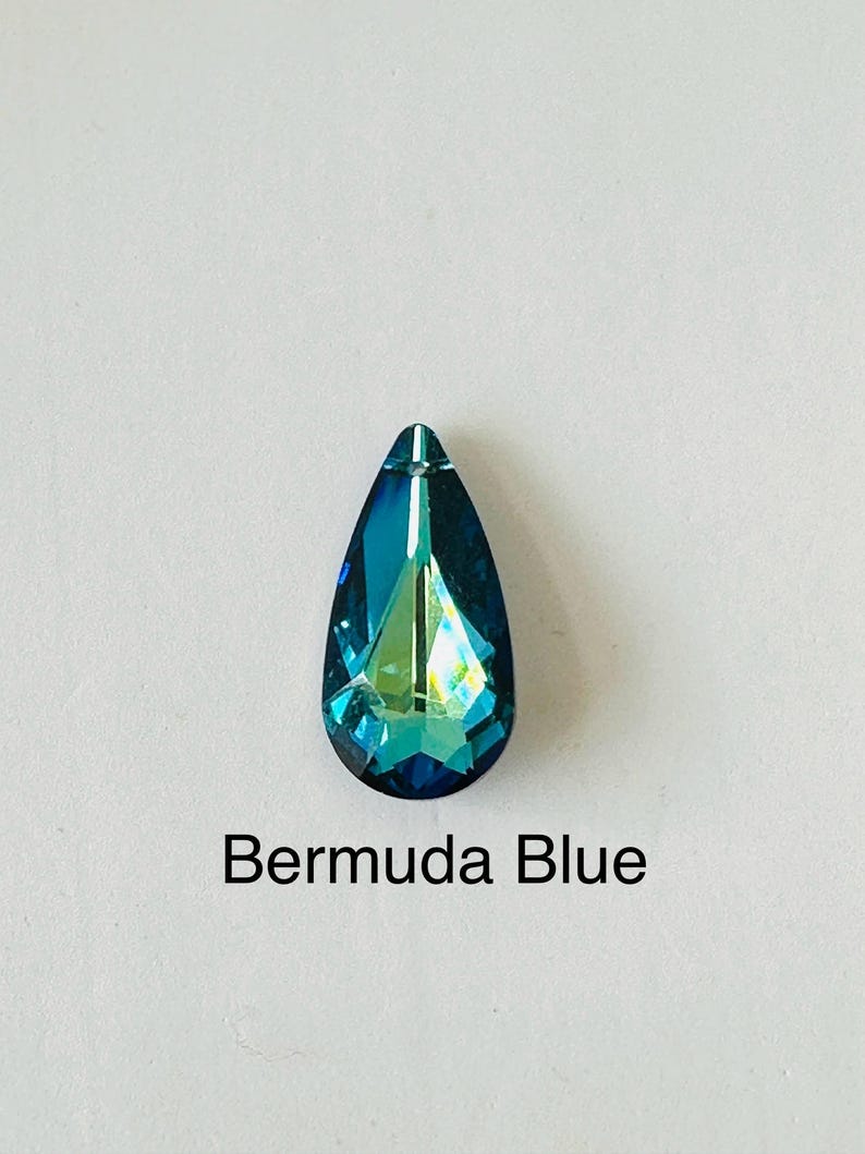 Op de afbeelding: Een druppelvormig Bermuda Blue kristal met een puntige top. Het kristal heeft een facetgeslepen oppervlak dat licht reflecteert, waardoor een glinsterend effect ontstaat. De tekst "Bermuda Blue" staat eronder.