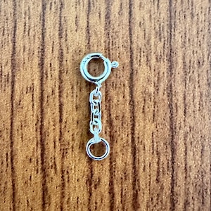 May include: A silver-colored necklace extender with a spring ring clasp and a small chain. The extender has a small jump ring at the end of the chain. The background is a wooden surface.