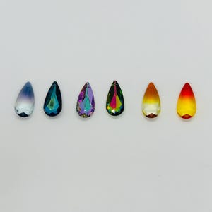 Op de afbeelding: Zes druppelvormige kristallen hangers op een rij, elk met een andere kleurovergang. De kleuren vari&euml;ren van blauw tot groen, paars en geel tot rood. De kristallen zijn facetgeslepen en reflecteren licht.