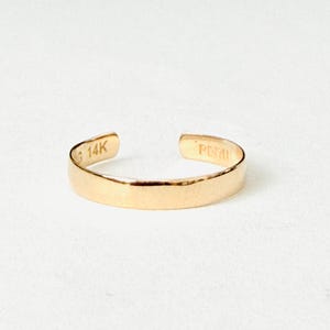 May include: A gold open-ended band ring with a smooth, polished finish. The interior of the band features the stamped text 14K and PERU. The ring has a simple, flat profile and a consistent width throughout its circular, adjustable design.