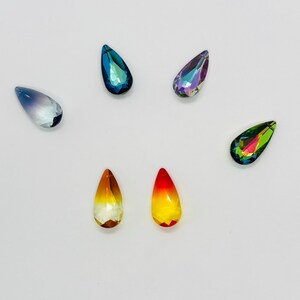 Op de afbeelding: Een verzameling druppelvormige kristallen strass-steentjes in verschillende kleuren. De strass-steentjes bevatten tinten blauw, paars, geel, oranje en groen. Elke steen heeft een facetgeslepen oppervlak, dat licht reflecteert en een sprankelend effect cre&euml;ert.