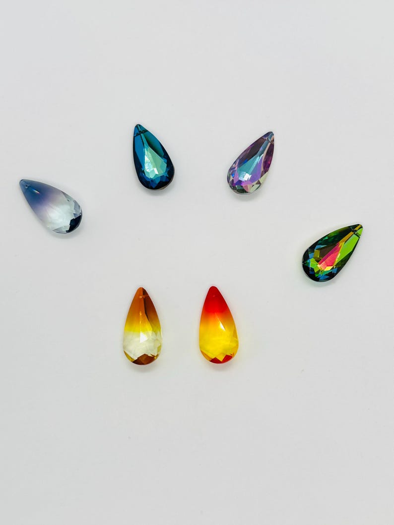 Op de afbeelding: Een verzameling druppelvormige kristallen strass-steentjes in verschillende kleuren. De strass-steentjes bevatten tinten blauw, paars, geel, oranje en groen. Elke steen heeft een facetgeslepen oppervlak, dat licht reflecteert en een sprankelend effect cre&euml;ert.