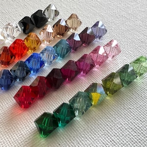 May include: Assortment of faceted glass beads in various colors, including clear, red, green, blue, purple, and black. The beads are arranged in rows on a white surface.