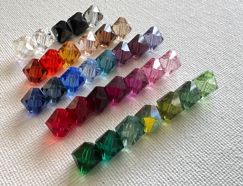 May include: Assortment of faceted glass beads in various colors, including clear, red, green, blue, purple, and black. The beads are arranged in rows on a white surface.