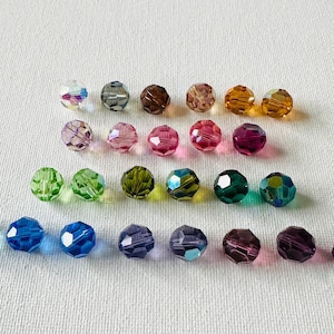 May include: An assortment of faceted beads in various colors, including blue, green, pink, purple, and amber. The beads are arranged in rows on a white surface, showcasing their different hues and textures. Ideal for jewelry making.