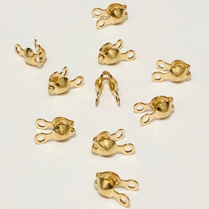 May include: A collection of small, gold-colored crimp end findings, designed for jewelry making. Each finding has a rounded body with two loops extending outwards. The findings are scattered on a white surface.
