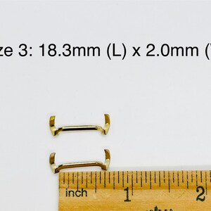 May include: Two gold-colored metal watch band spring bars, measuring 18.3mm in length and 2.0mm in width, are shown next to a ruler.