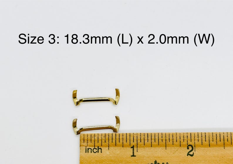 May include: Two gold-colored metal watch band spring bars, measuring 18.3mm in length and 2.0mm in width, are shown next to a ruler.