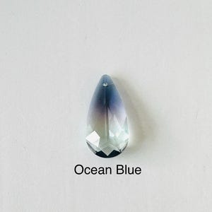 Op de afbeelding: Een gefacetteerde, druppelvormige kristallen hanger met een kleurengradient, die overloopt van diepblauw aan de bovenkant naar een helder, lichtblauw aan de onderkant. De tekst "Ocean Blue" is eronder gedrukt.