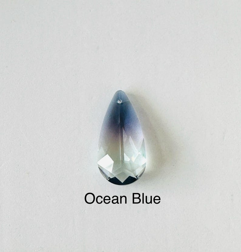 Op de afbeelding: Een gefacetteerde, druppelvormige kristallen hanger met een kleurengradient, die overloopt van diepblauw aan de bovenkant naar een helder, lichtblauw aan de onderkant. De tekst "Ocean Blue" is eronder gedrukt.