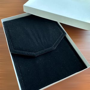 May include: An open white box with a black velvet interior. The interior features a flap with a geometric design. The box is on a brown surface.