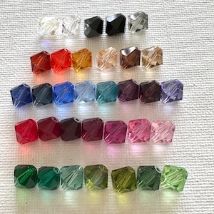 May include: Assortment of 20 square glass beads in various colors, including clear, red, blue, purple, green, and black. The beads are arranged in rows, with each row containing a different color.