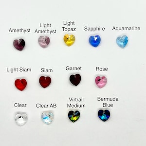 Puede incluir: Una colección de doce cristales en forma de corazón dispuestos en tres filas, etiquetados con los nombres de colores: Amethyst, Light Amethyst, Light Topaz, Sapphire, Aquamarine, Light Siam, Siam, Garnet, Rose, Clear, Clear AB, Virtrail Medium y Bermuda Blue.