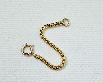 May include: A gold-toned box chain extender featuring a circular spring ring clasp on one end and a small jump ring on the other. The chain consists of interconnected square links that form a flexible, rounded, and continuous metal strand.