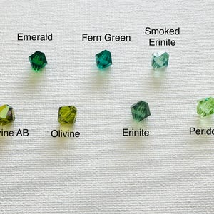 May include: A close-up of ten different shades of green glass beads. The beads are arranged in two rows of five. The beads are labeled with their names: Emerald, Fern Green, Smoked Erinite, Olivine AB, Olivine, Erinite, Peridot.