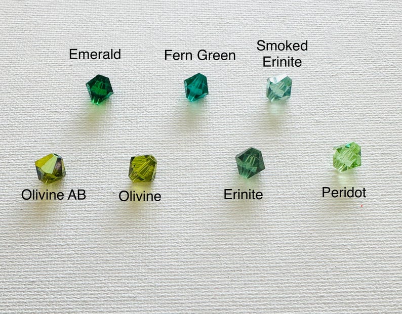 May include: A close-up of ten different shades of green glass beads. The beads are arranged in two rows of five. The beads are labeled with their names: Emerald, Fern Green, Smoked Erinite, Olivine AB, Olivine, Erinite, Peridot.