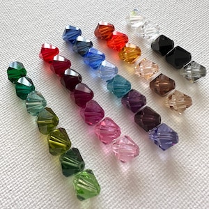 May include: Assortment of 12 faceted glass beads in various colors, including green, red, blue, purple, pink, and brown. The beads are arranged in rows on a white surface.
