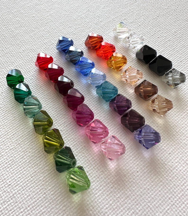 May include: Assortment of 12 faceted glass beads in various colors, including green, red, blue, purple, pink, and brown. The beads are arranged in rows on a white surface.